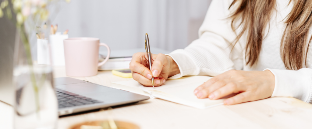 Woman writing with pen on piece of paper, while seated at desk. Her laptop is open on the desk in front of her and she has a pink mug of coffee to her right with a couple of white cups filled with an assortment of pens.